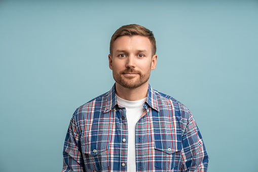 Portrait caucasian man with light smile on blue background looking at camera in plaid shirt. Studio shot bearded middle age friendly guy. White male feeling optimism. Enjoying life, sincere emotions.