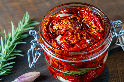 Sun-dried red tomatoes with garlic, green rosemary, olive oil and spices in a glass jar on a wooden table. Rustic style, close up