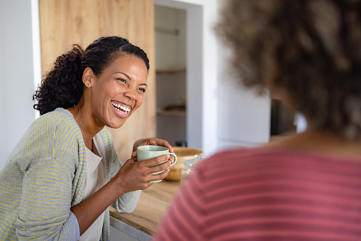 Cheerful African American woman drinking coffee and talking to her friend at home.