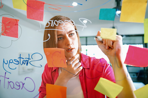 Closeup of young beautiful caucasian business leader presents marketing idea while writing marketing idea on glass board with mind map and colorful sticky notes. Portrait. Brainstorm. Immaculate.