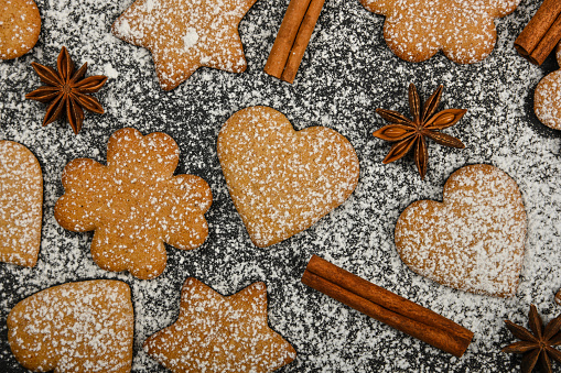 Close up heart and star shaped Christmas gingerbread cookies with cinnamon and star anise spices on black slate background with powdered white sugar icing, elevated top view, directly above