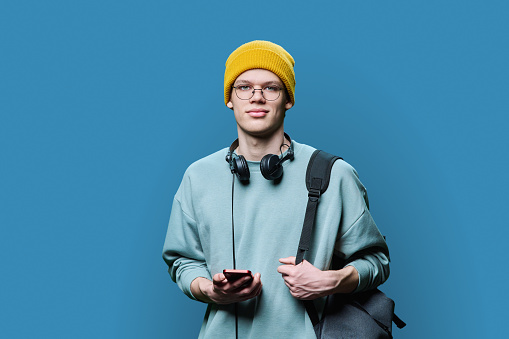 Young handsome male with headphones holding smartphone, blue background. Teenage guy college student looking at camera, hipster in knitted hat with backpack. Internet online technology, mobile apps