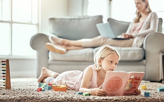 Girl child, home and reading book for learning, language development and mother work from home in living room. Family, kid and English story, school education and relax on floor with mom on sofa