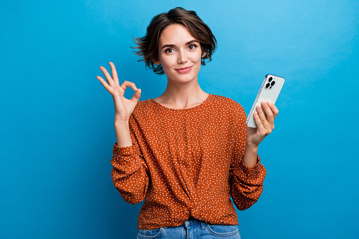 Photo portrait of attractive young woman hold device show okay symbol dressed stylish brown clothes isolated on blue color background.