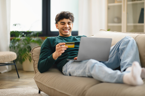 Online Shopping Offer. Happy Middle Eastern Young Man Holding Credit Card For Payment Using Laptop Computer Lying On Couch At Home. Great Sales And Ecommerce Concept. Selective Focus