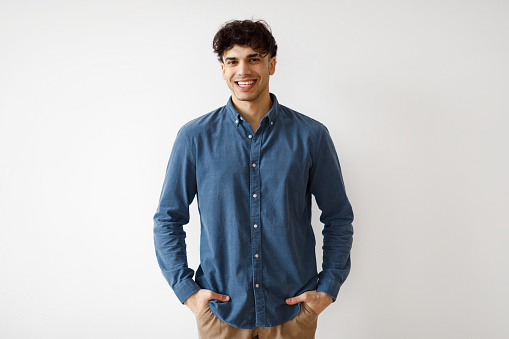 Happy Middle Eastern Young Man Smiling To Camera Standing Posing Near White Studio Wall Indoors. Shot Of Handsome Arabic Guy In Casual Clothing Expressing Positive Emotions