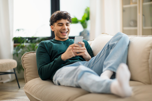 Happy Middle Eastern Young Man Using Cellphone And Laughing Texting Lying On Sofa At Home. Cheerful Man Holding Smartphone Playing Game And Networking Online Having Fun On Weekend
