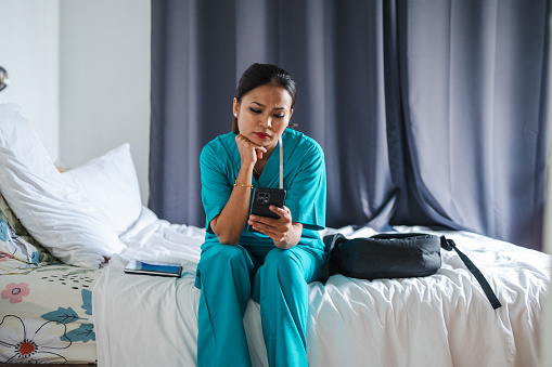 An Asian woman with scrubs sitting on her bed using smartphone. Lifestyle in scrubs