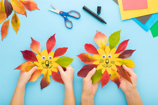 Mother and baby hands creating lion head shapes from colorful leaves and paper. Light blue table background. Making autumn decorations. Closeup. Playing and spending time together. Top down view.