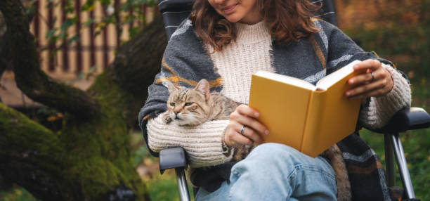 Young woman sitting in the autumn garden with a pet gray cat  reading a book. Leisure activity concept stock photo