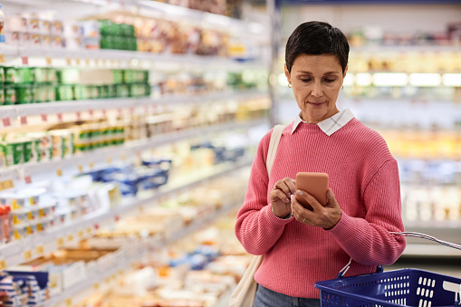 Waist up portrait of adult woman using smartphone in supermarket and holding basket, copy space
