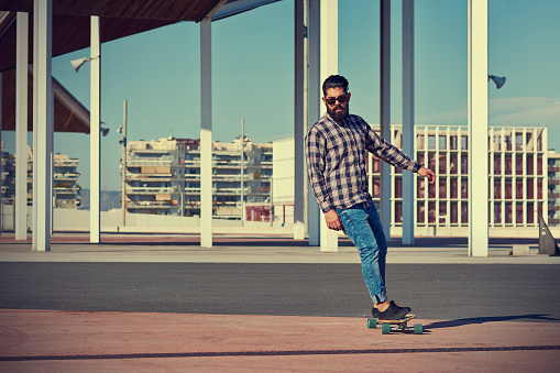 Full body of bearded male skater in checkered shirt and sunglasses riding longboard on paved road against buildings in sunlight