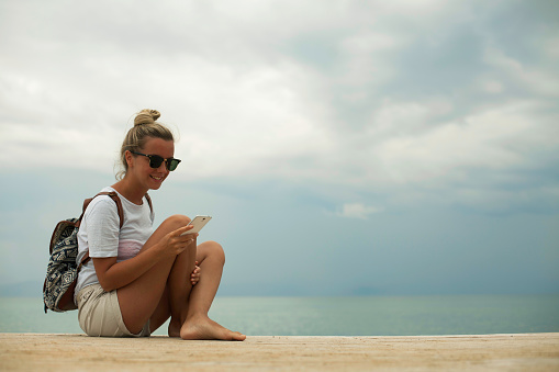 Young tourist woman is sitting on a pier and using mobile phone