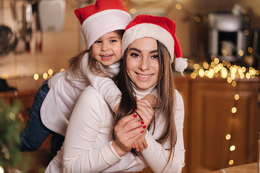 Portrait of adorable little daughter with her mom at kitchen during Christmas holidays. Girls in santa hat. Decorative fit tree at the table. Girls make gingerbread.