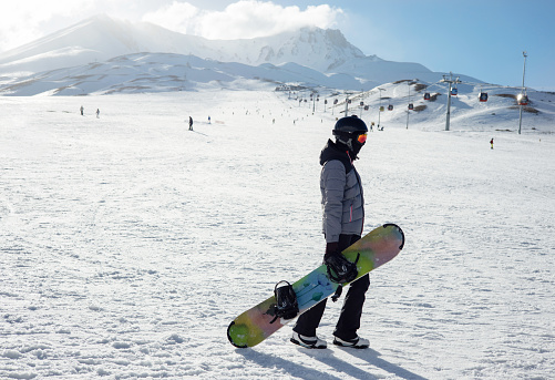 Girl or boy in ski helmet, sunscreen mask and balaclava with snowboard stands against the backdrop of snow-covered mountain ski slope and a cloudy sky. Winter. Extreme sport and travel content