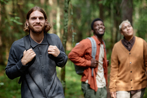 Portrait of young man with backpack looking at camera while walking in the forest together with his friends