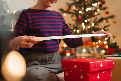 Close-up shot of woman wrapping presents for Christmas, sitting in living room decorated with a Christmas tree.