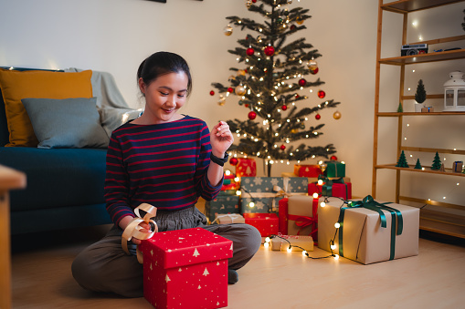Asian woman happily preparing to wrap a gift and having a new idea, sitting in living room decorated with a Christmas tree.