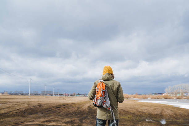 A man on a hike stands with a backpack on his shoulders with his back to the camera, a view from behind a man in a hat, a traveler against a cloudy sky. High quality photo