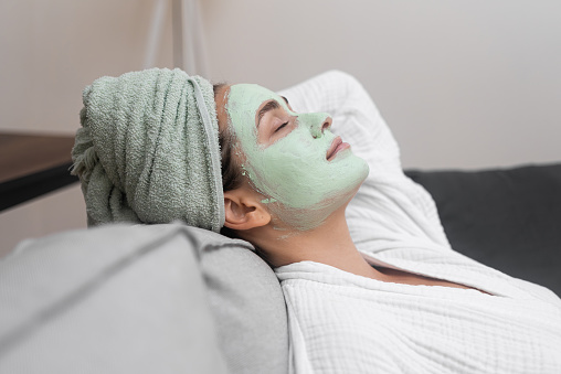 Sofa Serenity: A bathrobe-clad woman finds relaxation on a cozy sofa with a facial moisturizing mask, immersing herself in soothing home spa procedures.