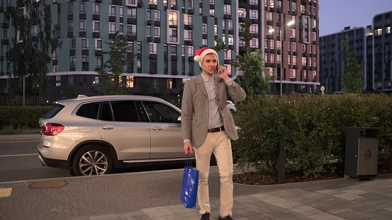 Happy young man in Santa Claus hat walking along the street, talking on mobile phone, car on the background. Business, celebration, New Year concept. Slow motion