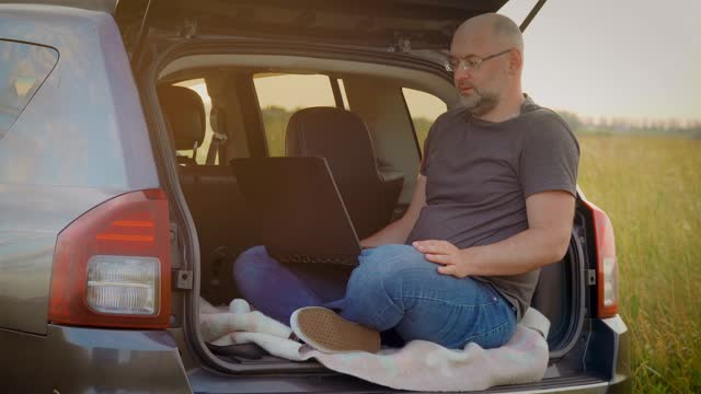 Adult man communicating by video call, speaking looking at laptop computer, online conference distance office chat, sitting in car trunk while traveling