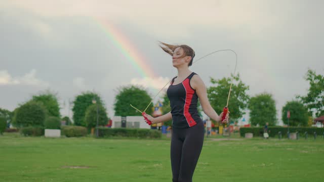 Young woman working on her fitness, skipping with jump rope against background of beautiful rainbow