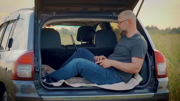 Busy bald adult man working remotely at laptop computer as sitting in car trunk while travelings