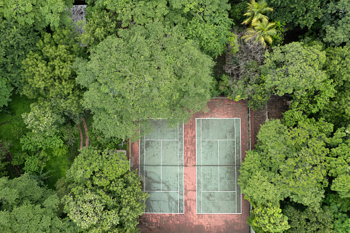 Aerial photograph captures  red clay tennis court nestled in the midst of a dense forest. The court is marked with white lines and surrounded by tall, lush trees that provide shade for the players.