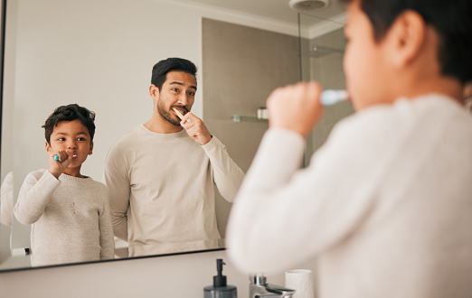 Dad, boy and child brushing teeth in mirror for hygiene, morning routine and teaching healthy oral habits at home. Father, kid and dental cleaning in bathroom with toothbrush, fresh breath and care