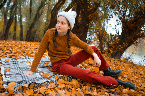 a young teenage girl is sitting in a glade by the river bank in an autumn forest, enjoying the beautiful nature and bright yellow leaves