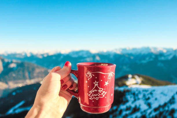 A woman's hand with a red mug with a Christmas pattern against the backdrop of a snowy mountain landscape. Background for holiday-themed content about Christmas and New Year.