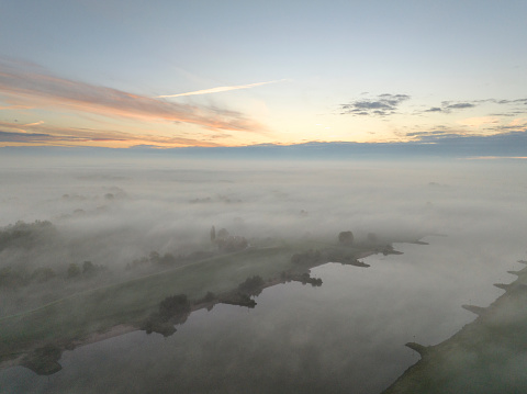 River IJssel covered in a blanket of mist seen from above during a fall morning near the river IJssel in Overijssel. The treetops are popping out over the thick fog with some of the trees starting to change color during the annual autumn season. The fog is giving the landscape a mystical atmosphere.