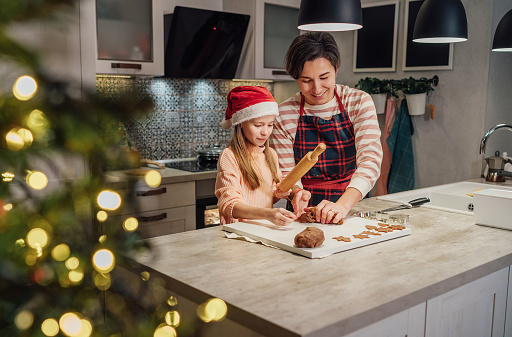 Cute little girl in red Santa hat with mother making homemade Christmas gingerbread cookies using rolling pin together in home kitchen. Happy family holidays preparation and childhood concept.