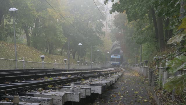 The funicular car arrives at the lower station. Railroad tracks in the foreground, blue cable car moves. Foggy autumn morning in park with colorful red and orange trees in the background