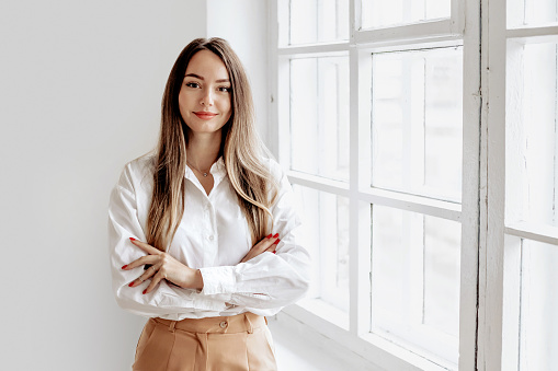 Portrait of a Caucasian business lady in a white shirt with folded hands against the background of an office window