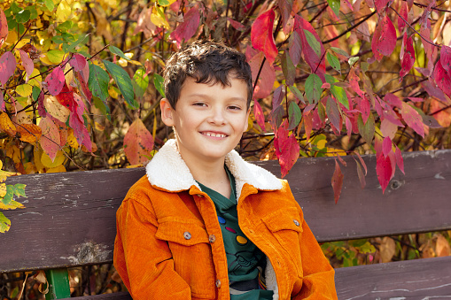 Charming boy in orange jacket is sitting on a bench in the park among bright autumn foliage.
