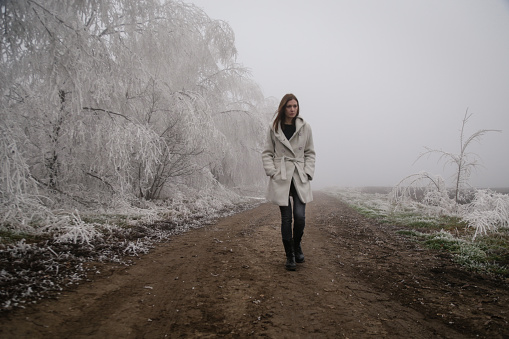 Woman in white coat walking on remote countryside rural road on cold freezing foggy day. Winter weather.