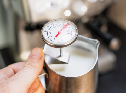 Close-up as a man holds up a jug of steamed milk with a temperature gauge, with an espresso coffee machine.