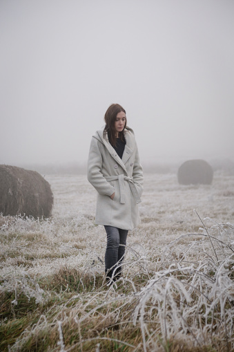 Woman in white coat walking through snowy countryside field on cold freezing foggy day.