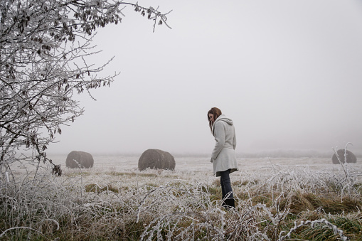 Woman in white coat walking through snowy countryside field on cold freezing foggy day.