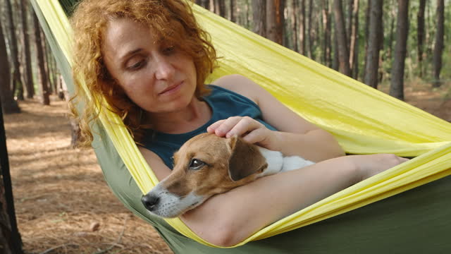 Concept Love for animals, friendship, happiness. Portrait red-haired woman dog close up camping in hammock relaxing in nature hugging dog summer pine forest. Caring for pets. Travel.