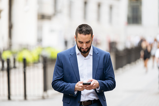 Front view of bearded mid adult wearing navy blue jacket over white open-collar shirt, walking outdoors and text messaging.