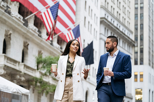 Business partners in office attire walking with takeaway coffee cups in Lower Manhattan, exchanging ideas, landmark financial building in background.