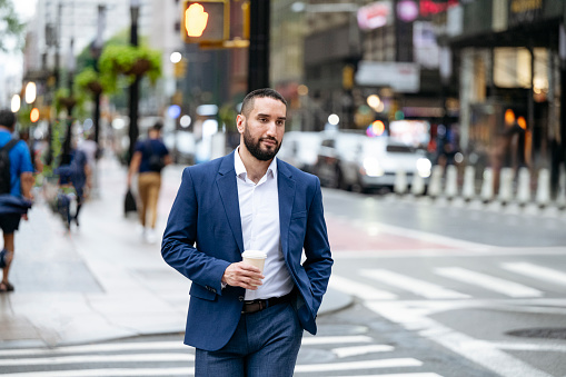 Bearded New York City office worker in early 30s crossing street with hand in pocket, holding takeaway cup.