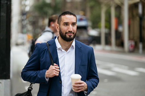 Waist-up view of early 30s bearded professional carrying gym bag over shoulder, holding takeaway coffee cup and crossing street.