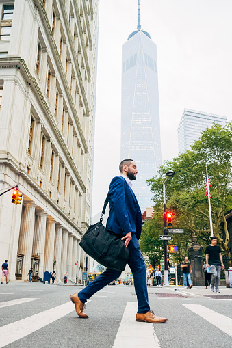 Full length side view of professional in blue suit, gym bag on shoulder, holding takeaway cup and walking home after work, One World Trade Center in background.