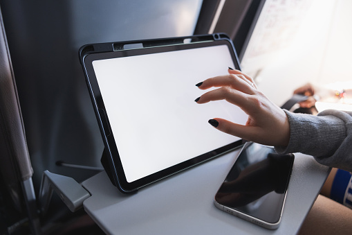 Young woman using a tablet and smartphone on an airplane with blank white desktop screen. Young female traveler using tablet pc while waiting her take off on aircraft, inside airplane cabin. Travel concept.