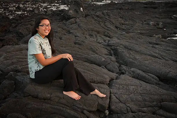 Barefoot young lady happily sitting on the seaside lavarock at sunset in Hawaii.
