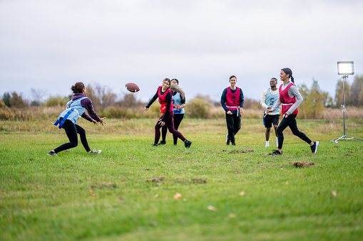 A group of University students run around on a field as they play a game of flag football together.  They are each dressed comfortably and have numbered team pinnies on.